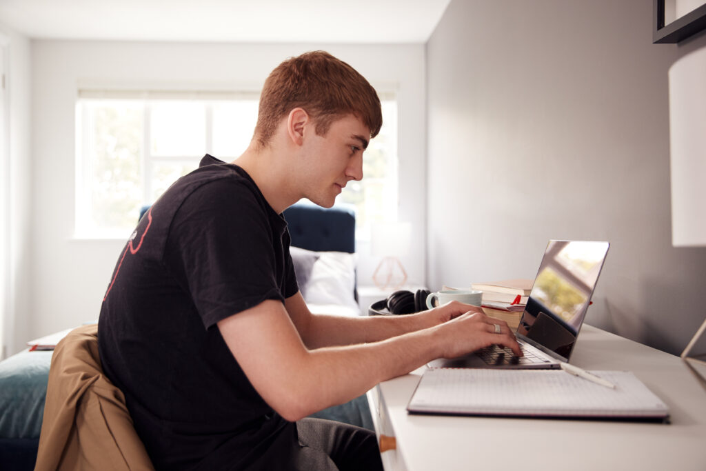 Male College Student In Shared House Bedroom Studying Sitting At Desk